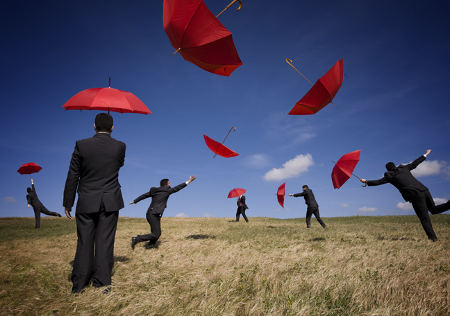businessmen running after red umbrellas in open grassy field