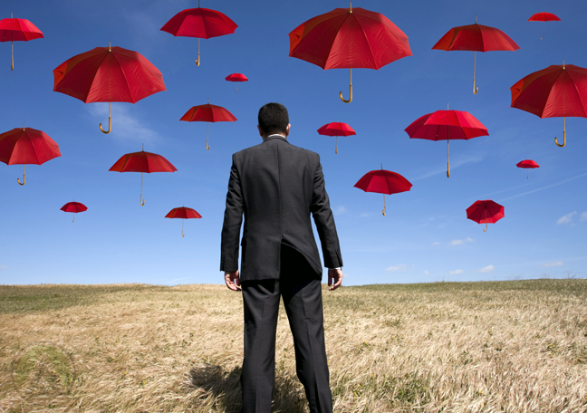 businessman-in-suite-standing-in-middle-of-the-field-looking-up-at-blue-sky-with-floating-red-umbrellas