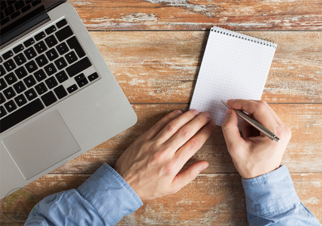 businessman-writing-with-pen-on-notepad-paper-next-to-laptop