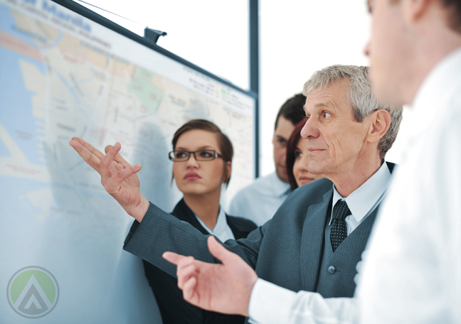 middle aged businessman pointing to map flanked by younger business team