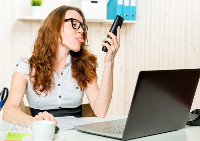 female office employee in glasses using laptop sticking tongue out at smartphone